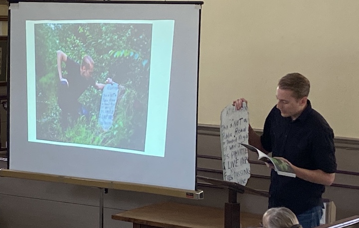 Presentation in Old Courthouse Museum showing a projected image on screen at left with Steve Skafte standing behind podium on the right holding prperty sign discovered on one of his journeys.