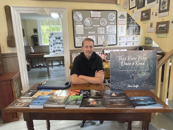 Steve Skafte sitting behind a table in the Old Courthouse Museum with all his books laid out on the table for purchase .